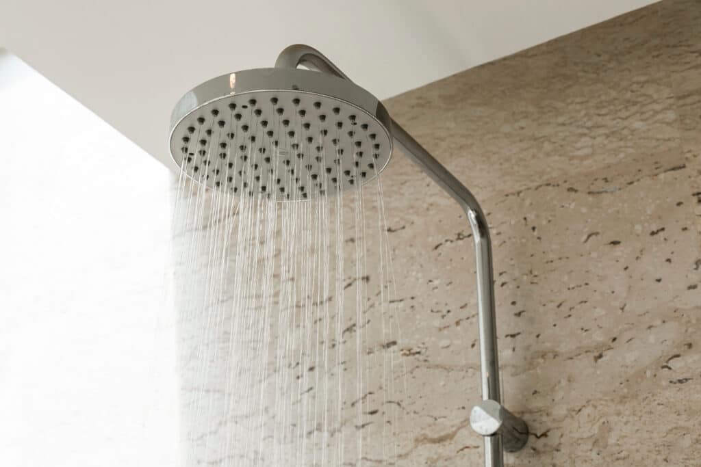 Grande pomme de douche ronde chromée en fonctionnement, jets d'eau fins tombant devant un mur de travertin beige.