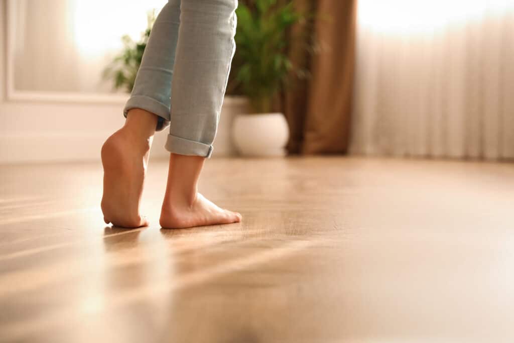 Pieds nus d'une personne marchant sur un sol en bois clair réchauffé par la lumière du soleil entrant par une fenêtre.