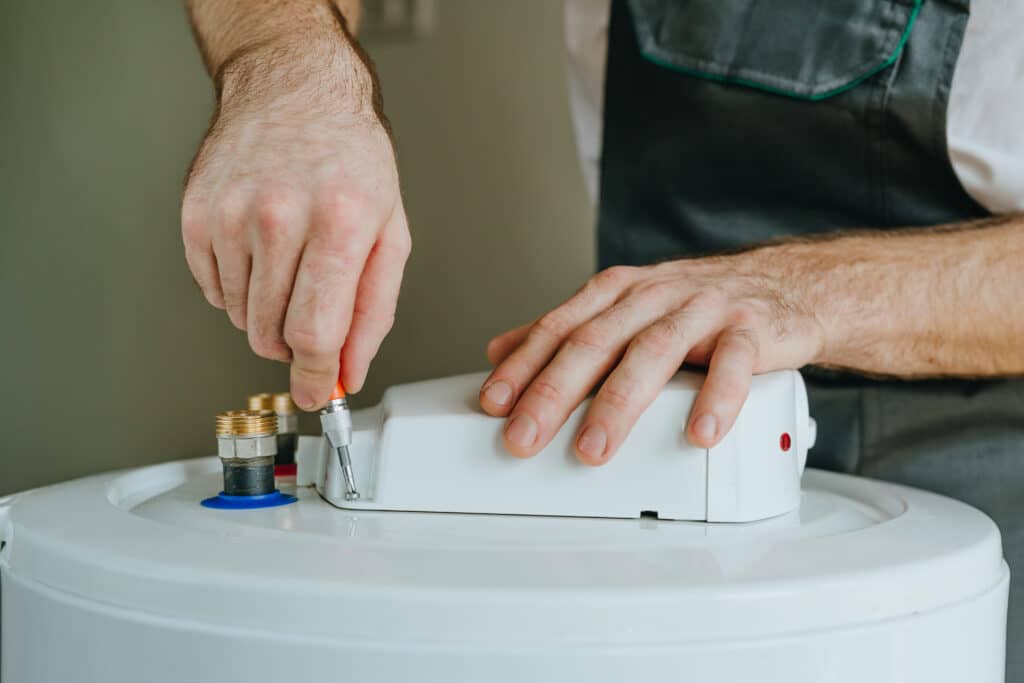 Un plombier utilise un tournevis pour ouvrir le boîtier d'un chauffe-eau électrique blanc, près des raccords en laiton.