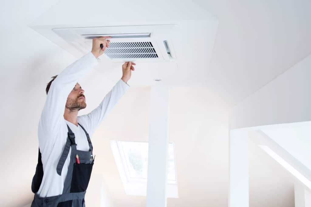 Technicien en combinaison vérifie un climatiseur encastré au plafond dans un grenier lumineux avec puits de lumière.